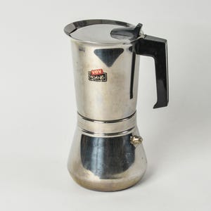 May include: A stainless steel stovetop espresso maker with a black handle and lid. The coffee maker has a classic design, with a cylindrical body and a conical base. The brand name "vev vagano" is visible on the front.