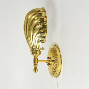 May include: A brass wall sconce with a shell-shaped shade and a round backplate. The sconce has a detailed, ornate design and is mounted on a white wall. The shade is a bright gold color.