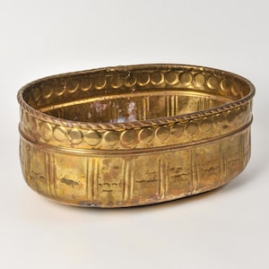 May include: An oval brass planter featuring a decorative rope-style rim and a band of embossed circles along the upper edge. The exterior body displays vertical fluted panels and a weathered, golden metallic finish with visible signs of patina.