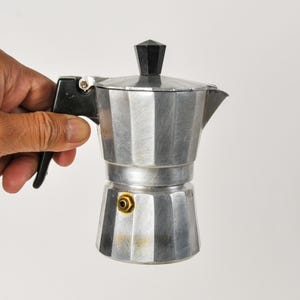 May include: A silver-colored aluminum moka pot with a faceted octagonal body, a black plastic handle, and a black knob on the lid. The base features a small brass-colored pressure release valve on the side, and the metal surface shows a brushed finish.