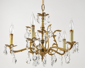 May include: A gold-toned metal chandelier featuring four candle-style light sockets with cream-colored sleeves. The fixture is adorned with numerous clear, teardrop-shaped glass crystals hanging from ornate, curved arms and a central column.