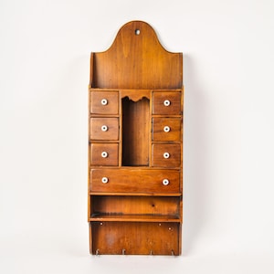 May include: A wall-mounted wooden organizer featuring a warm brown finish, six small drawers with white knobs, one wide drawer, an open central compartment, a lower shelf, and four metal hooks at the bottom for hanging items.