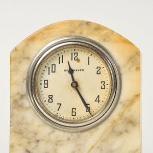 K&ouml;nnte beinhalten: Eine bogenf&ouml;rmige Kaminuhr mit cremefarbenem Zifferblatt, schwarzen arabischen Ziffern und dem Schriftzug New Haven. Die Uhr hat schwarze Zeiger und ist in eine silberfarbene Metalll&uuml;nette eingefasst, die in einem glatten, beigen Marmorgeh&auml;use sitzt.