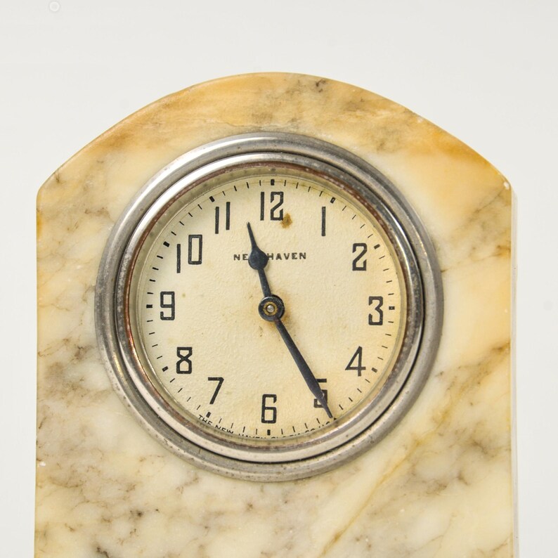 K&ouml;nnte beinhalten: Eine bogenf&ouml;rmige Kaminuhr mit cremefarbenem Zifferblatt, schwarzen arabischen Ziffern und dem Schriftzug New Haven. Die Uhr hat schwarze Zeiger und ist in eine silberfarbene Metalll&uuml;nette eingefasst, die in einem glatten, beigen Marmorgeh&auml;use sitzt.