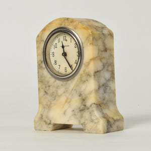 K&ouml;nnte beinhalten: Eine Kaminuhr mit einem Geh&auml;use aus cremefarbenem und grauem Marmorstein, das eine gew&ouml;lbte Oberseite und ein rundes Zifferblatt aufweist. Das Zifferblatt ist wei&szlig; mit schwarzen arabischen Ziffern und feinen Zeigern, eingefasst in einen silberfarbenen Metallrahmen.