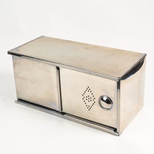 May include: A rectangular, stainless steel bread box with two sliding doors. The box has a diamond-shaped ventilation pattern and a round handle on the right door. The box is made of shiny, reflective metal.