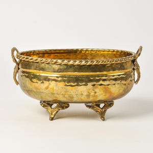 May include: An oval brass bowl featuring a hammered texture on the exterior, a rope-style rim, and two decorative handles on the sides. The bowl is supported by three ornate, leaf-shaped feet and has a polished, metallic gold-toned finish.