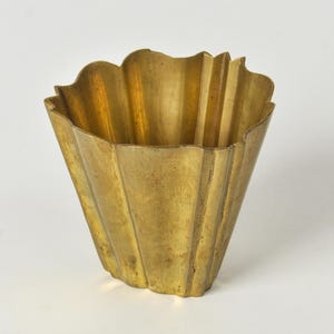 May include: A gold-colored, fluted metal vase with a scalloped rim. The vase has a wide, open top and a tapered base. The surface has a slightly aged appearance, with vertical lines adding texture. The design is reminiscent of Art Deco.