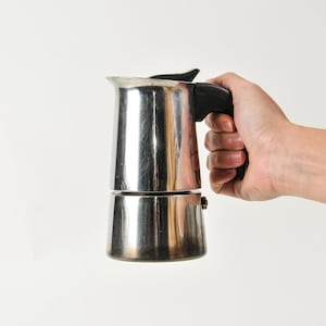 May include: A stainless steel stovetop espresso maker with a black handle and lid. The coffee pot is held by a hand against a white background. The pot is designed to brew coffee on a stovetop.
