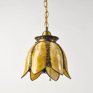 May include: A hanging pendant lamp featuring a bell-shaped shade made of amber and cream swirled glass panels. The shade is accented with ornate bronze-toned metal filigree trim along the edges and hangs from a matching bronze chain.