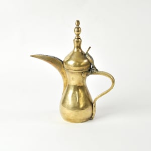 Puede incluir: Una cafetera de latón antigua con un pico largo y un asa curvada. La cafetera tiene un cuerpo bulboso y un adorno decorativo en la tapa. El latón tiene un tono dorado cálido y parece bien usado.