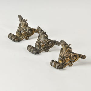 May include: Three matching metal furniture feet featuring an ornate, sculpted face design with a dark, aged bronze finish. Each piece displays intricate scrollwork and leaf-like detailing, with a protruding metal pin for attachment purposes.