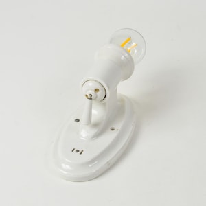 May include: A white ceramic wall sconce with a clear lightbulb. The sconce has a rounded base and a cylindrical arm that holds the bulb. The lightbulb has a yellow filament. The sconce is designed to be mounted on a wall.