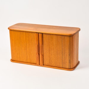 May include: A rectangular wooden storage cabinet featuring a warm brown grain and two sliding tambour doors. The cabinet has a smooth top surface with rounded edges and vertical slats that form the front panels, each with a vertical handle.
