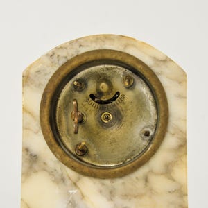 K&ouml;nnte beinhalten: Die R&uuml;ckseite einer Uhr mit einem Geh&auml;use aus wei&szlig;-grauem Marmorstein. In der Mitte ist eine kreisf&ouml;rmige Messingplatte eingelassen, die einen kleinen Aufziehschl&uuml;ssel, drei Metallstifte und eine geschwungene &Ouml;ffnung mit dem Text Fairfield enth&auml;lt.