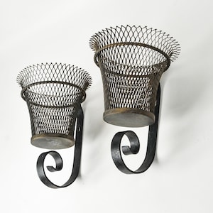 May include: Two antique metal wall planters. Each planter features a woven wire basket with a decorative rim, mounted on a curved, wrought iron bracket. The baskets have a bronze-colored base and rim. The brackets are black.