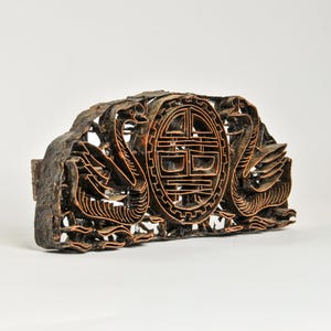 May include: A dark brown carved wooden printing block featuring two symmetrical swan figures facing a central oval medallion. The medallion contains intricate geometric lines and symbols. The wood surface shows a weathered texture.