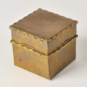 May include: A square brass box with a weathered, metallic finish. The lid and the base feature a decorative border that mimics the segmented appearance of bamboo stalks. The box has a flat top and straight sides, showing a consistent golden-brown tone.
