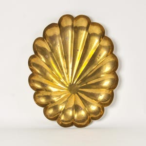 May include: A polished brass dish shaped like a large scallop shell. The metal object features a series of deep, radiating fluted grooves that extend from the center to the scalloped outer edge, creating a textured, reflective golden surface.