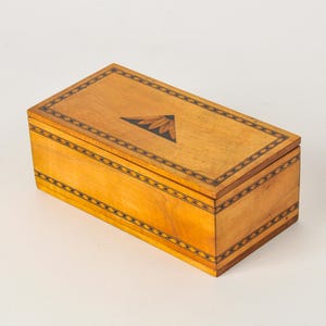 May include: A rectangular wooden box with a light brown finish, featuring a dark brown geometric border pattern along the edges of the lid and sides. The center of the lid displays a dark triangular inlay with a stylized floral petal design.