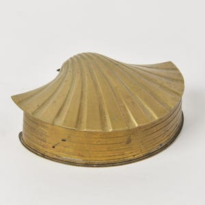 May include: A brass trinket box shaped like a scallop shell. The lid features a radial ribbed texture, while the base has a smooth, curved side profile. The metal has a warm, golden-yellow tone with subtle variations in the polished surface finish.