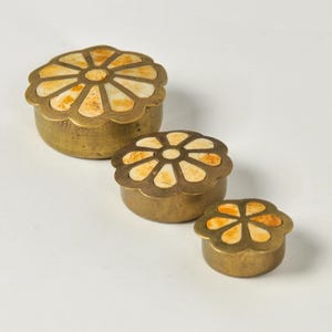 May include: A set of three round brass containers with lids, each featuring a scalloped edge and an inlaid floral pattern made of cream and tan mottled material. The containers are arranged in a descending size order from largest to smallest.