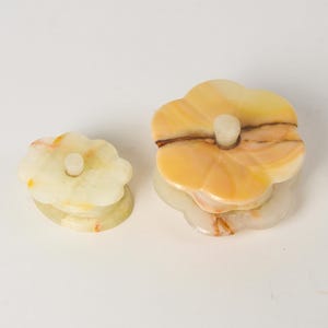 May include: Two flower-shaped stone trinket boxes with lids. The smaller box is pale cream with subtle yellow veining. The larger box is a warm golden yellow with darker brown mineral streaks. Each lid features a small, cylindrical stone knob.