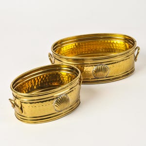 May include: Two oval-shaped brass containers with a hammered texture and a polished gold finish. Each vessel features small side handles and a prominent embossed seashell motif on the front exterior, showcasing a reflective metallic surface.