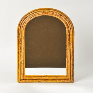 May include: An arched mirror with a natural bamboo frame. The frame is made of multiple layers of bamboo, creating a textured, layered effect. The mirror has a rectangular shape with a rounded top. The mirror's surface is a dark brown color.