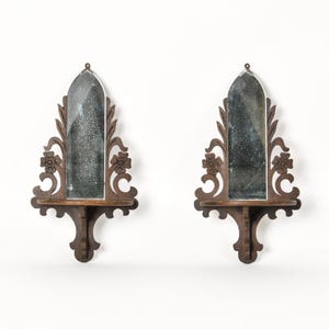 May include: A pair of dark brown wooden wall shelves, each featuring an arched mirror back panel with a weathered finish. The wooden frames are intricately carved with floral and scrollwork details, including a small shelf base.