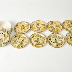 May include: A set of eight decorative coasters and a round box. The coasters and box lid feature a cream-colored background with a pattern of yellow and black butterflies. The box is open, revealing its interior.