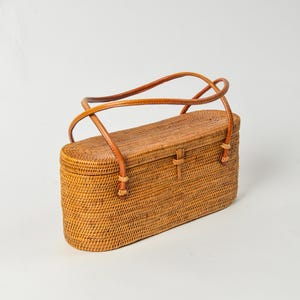 May include: A rectangular, woven rattan handbag with a light brown leather handle. The bag has a clasp closure and decorative accents at the handle attachments. The bag is a natural tan color and appears to be handmade.