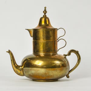 May include: A multi-tiered brass teapot featuring a wide, rounded base with a curved spout and handle, topped with a smaller cylindrical section and a pointed lid. The metal surface displays a polished gold-toned finish with subtle variations.