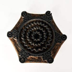 May include: A hexagonal wooden block stamp featuring a dark brown finish and a raised black carved design. The pattern consists of a central star, concentric zigzag rings, and floral motifs at each of the six corners of the wooden block.