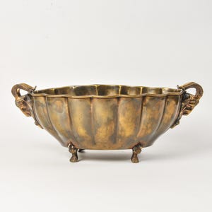 May include: An oval brass bowl with a scalloped edge and a warm, aged patina. The bowl features two handles shaped like ram heads on either side and is supported by four small, detailed feet at the base, creating a distinct, elevated profile.