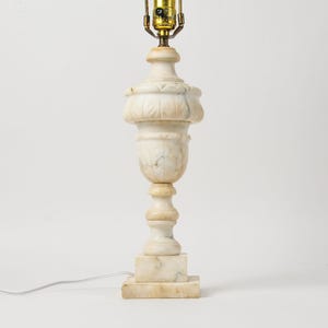 May include: A table lamp base crafted from off-white and beige veined marble. The lamp features a central urn-shaped body with carved leaf detailing, a tiered pedestal base, and a gold-toned metal socket assembly at the top with a cord.