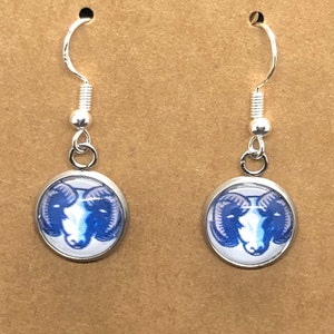 May include: A pair of silver-toned dangle earrings. Each earring features a round, silver-toned bezel with a blue and white ram's head design. The earrings have a curved hook for pierced ears.