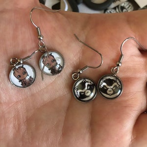 May include: A pair of silver dangle earrings with round glass charms. One charm features a cartoon image of Groot from Guardians of the Galaxy. The other charm features a cartoon image of Rocket Raccoon from Guardians of the Galaxy.