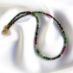 May include: A green and pink gemstone necklace with gold beads. The necklace is made of small, round beads and has a clasp closure.