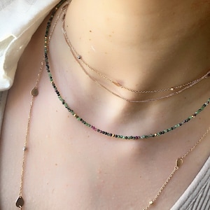 May include: A layered necklace set featuring a delicate gold chain with a small pendant, a thicker gold chain, and a beaded chain with green, black, and pink gemstones.
