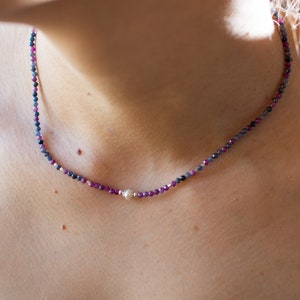 May include: Delicate necklace featuring small, faceted beads in shades of deep red, purple, and blue. A single, round, silver-colored bead is centered on the necklace. The necklace is designed to be worn close to the neck.