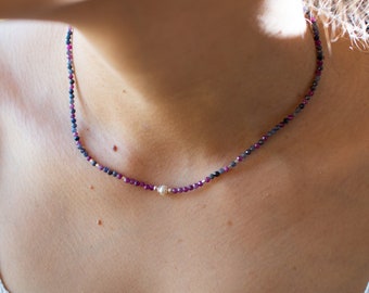 May include: Delicate necklace featuring small, faceted beads in shades of deep red, purple, and blue. A single, round, silver-colored bead is centered on the necklace. The necklace is designed to be worn close to the neck.
