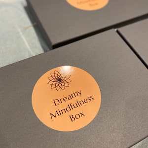 以下が含まれることがあります： 「Dreamy Mindfulness Box」と書かれた丸いオレンジ色のステッカーが貼られた灰色の箱。ステッカーには、スタイリッシュな花柄のデザインが施されています。