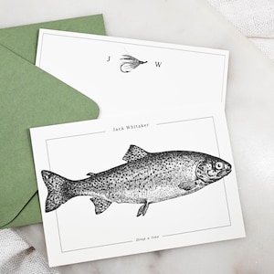 以下が含まれることがあります： 魚とフライフィッシングの毛鉤の黒いインクイラストが描かれた白いステーショナリーカード。1枚のカードにはJack Whitakerという名前とDrop a lineというテキストがあり、もう1枚にはJ Wのイニシャルが記されています。セージグリーンの封筒が付属しています。
