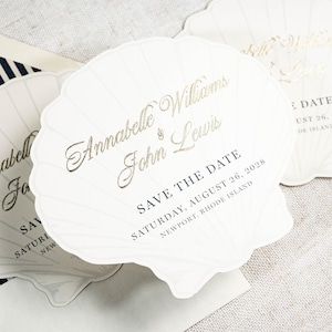 Puede incluir: Tres tarjetas blancas con forma de concha con letras en lámina dorada que dicen Annabelle Williams and John Lewis, SAVE THE DATE, SATURDAY, AUGUST 26, 2028, NEWPORT, RHODE ISLAND. Las tarjetas tienen estrías en relieve y un sobre a rayas.