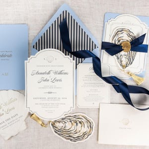 May include: A wedding stationery set featuring light blue and cream cards with navy blue ribbon, gold wax seals, and oyster shell illustrations. The text includes Annabelle Williams and John Lewis, Saturday, August 24, 2024, and We can't wait to celebrate with you.