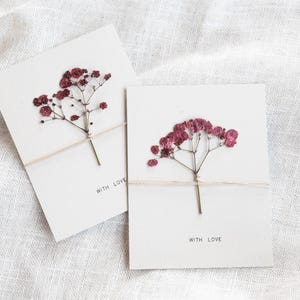 May include: Two white cards, each adorned with dried burgundy flowers and tied with twine. The cards are printed with the words "WITH LOVE". They rest on a white linen background.