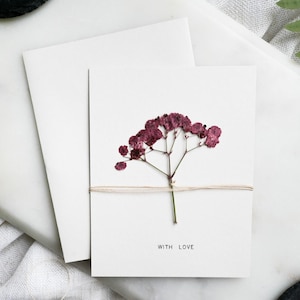 Puede incluir: Tarjeta de felicitación blanca con una flor burdeos seca y la frase «WITH LOVE». La tarjeta está atada con hilo y descansa sobre un sobre blanco. Un helecho y un posavasos negro están en el fondo.