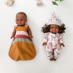May include: Two Black baby dolls wearing different outfits. One doll is wearing a brown and pink striped sleep sack with a brown bottom. The other doll is wearing a pink and white onesie with a deer print. The dolls are on a white surface with a rainbow yarn wall hanging and a potted plant in the background.