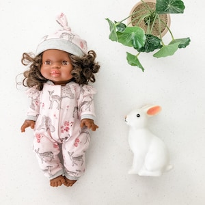 May include: A pink and grey doll wearing a grey and pink deer print onesie and a grey and pink knotted hat. The doll has brown eyes and brown curly hair. A white rabbit and a potted green plant are in the background.